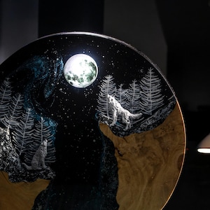 Puede incluir: Obra de arte redonda de madera que representa una escena nocturna con una luna llena y dos lobos aullando. El fondo es azul oscuro con estrellas y &aacute;rboles blancos. Los lobos son de color gris claro.