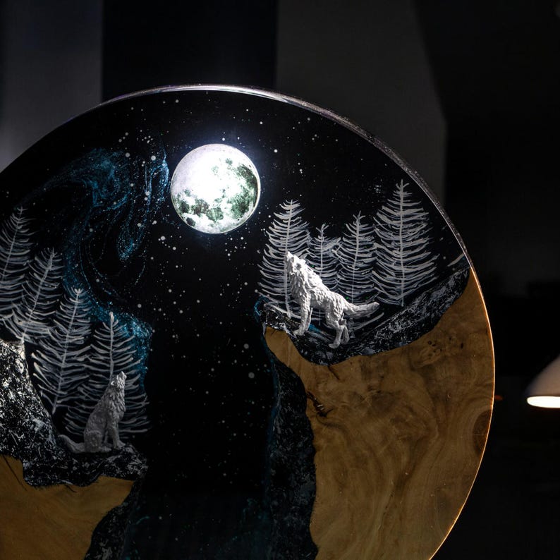 Puede incluir: Obra de arte redonda de madera que representa una escena nocturna con una luna llena y dos lobos aullando. El fondo es azul oscuro con estrellas y &aacute;rboles blancos. Los lobos son de color gris claro.