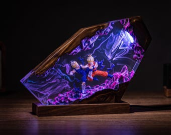 Puede incluir: Una lámpara personalizada de resina y madera con dos figuras de acción de anime en pose de combate. La resina transparente contiene efectos de humo violeta y azul, iluminados por una luz interna, dentro de un marco de madera oscura.