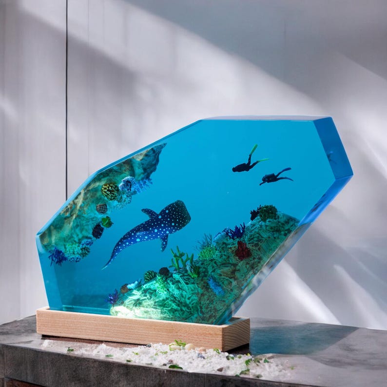 May include: A blue resin sculpture of a whale shark swimming in a coral reef with two scuba divers. The sculpture is on a wooden base.