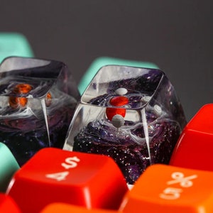 May include: Close-up of three custom keyboard keycaps with a galaxy-like design. The keycaps are made of clear resin with swirls of purple, white, and black. One keycap has a red accent. The keycaps are sitting on a keyboard with red and orange keys.
