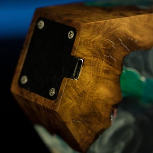 May include: Close-up of a wooden block with a dark, textured finish. A black metal plate is attached with silver screws. A metal port is visible on the side. The wood grain is prominent, with a natural, warm color.