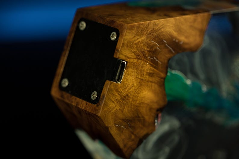 May include: Close-up of a wooden block with a dark, textured finish. A black metal plate is attached with silver screws. A metal port is visible on the side. The wood grain is prominent, with a natural, warm color.