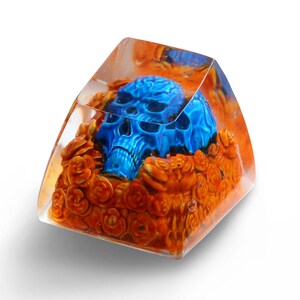 May include: A blue skull resin keycap with orange floral details encased in clear resin.
