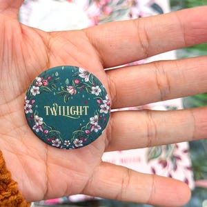May include: A round, teal button with the word "TWILIGHT" in gold script. The button is decorated with a floral wreath of white and pink flowers. The button is held in a person's hand.