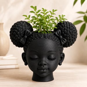 以下が含まれることがあります： つや消しの黒い頭の形をしたプランターで、彫刻された顔、閉じた目、ふっくらとした唇が特徴です。髪は2つの大きなテクスチャーのあるアフロパフにスタイリングされ、それぞれに小さなリボンが飾られています。頭の上部から小さな緑の植物が生えています。