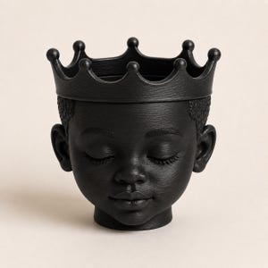 May include: A matte black planter shaped like the head of a young person with closed eyes and textured hair, topped with a crown featuring eight rounded points. The surface has a subtle horizontal grain texture throughout the entire object.