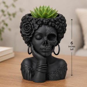 May include: A black planter shaped like a person with a half-face skull design, curly hair, and a rose accent. The planter holds a green succulent and measures 6 inches tall. The figure wears hoop earrings and bracelets, with a decorative neckline.
