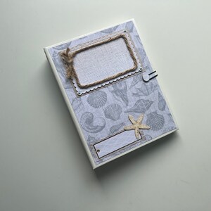 Puede incluir: Un &aacute;lbum de recortes blanco con un patr&oacute;n de conchas marinas azul claro y gris. La portada tiene un marco de cuerda marr&oacute;n con una etiqueta blanca y una peque&ntilde;a estrella de mar.