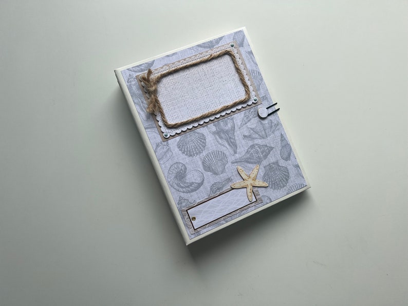 Puede incluir: Un &aacute;lbum de recortes blanco con un patr&oacute;n de conchas marinas azul claro y gris. La portada tiene un marco de cuerda marr&oacute;n con una etiqueta blanca y una peque&ntilde;a estrella de mar.