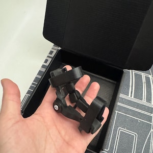 May include: A black, articulated, multi-jointed plastic mount featuring two adjustable fabric straps. The device has a central hinge secured with a silver-colored metal screw and is designed with a rigid frame structure for mounting purposes.