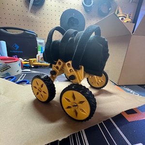 May include: A gold-colored, four-wheeled robot with black tires and a black fabric seat. The robot is on a brown paper surface.