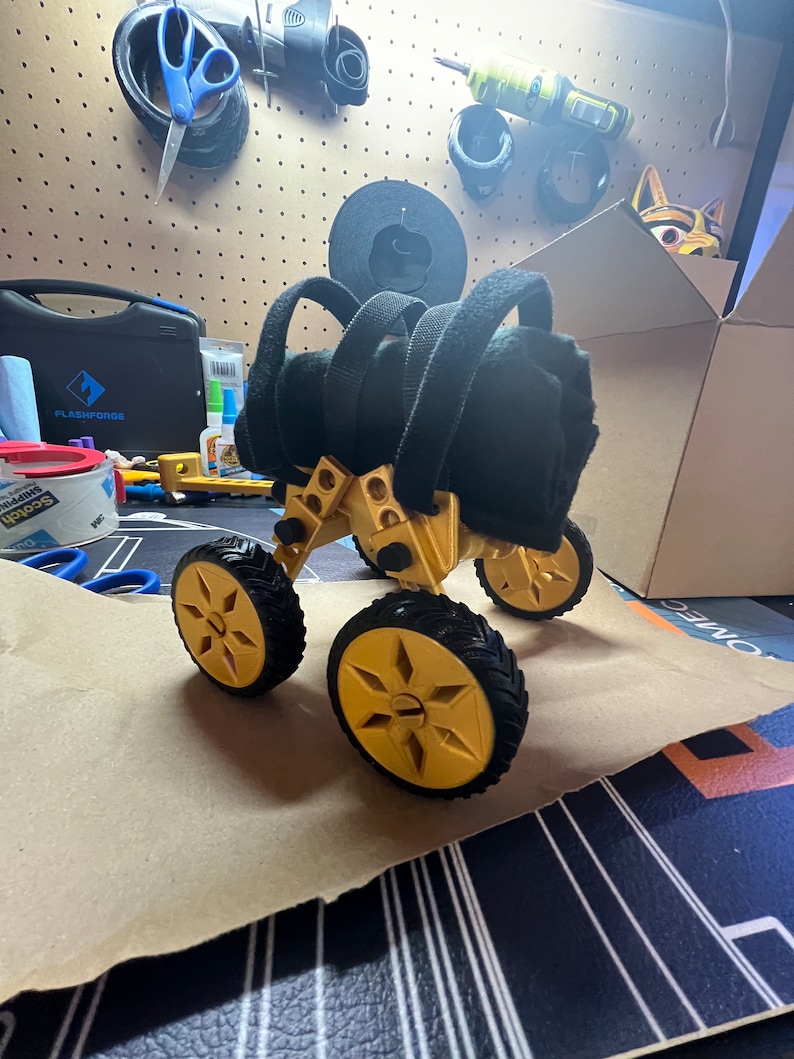 May include: A gold-colored, four-wheeled robot with black tires and a black fabric seat. The robot is on a brown paper surface.