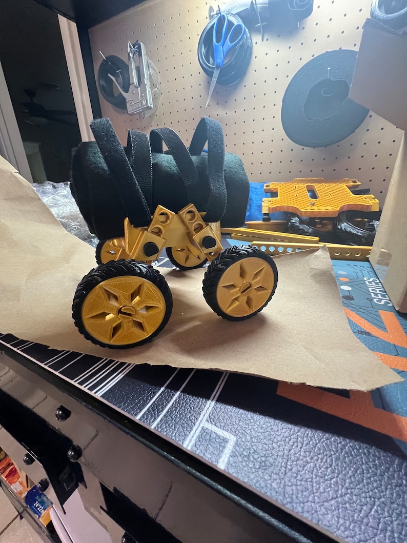 May include: A gold-colored toy vehicle with four wheels and black straps. The vehicle is made of plastic and has a Lego-like design.