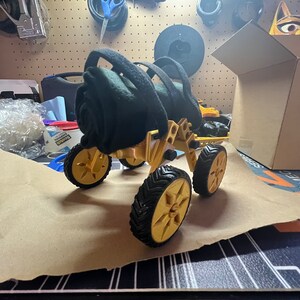 May include: A gold-colored, four-wheeled, all-terrain vehicle with black tires. The vehicle has a black fabric roll attached to the top.