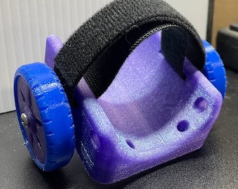 May include: A purple 3D printed device with a black velcro strap and a blue wheel. The device has a textured surface and several small holes.