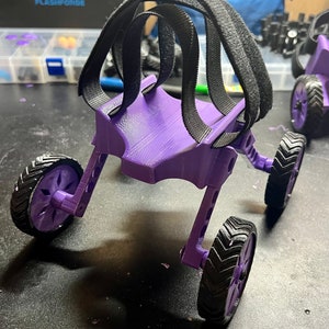 May include: A purple 3D printed, four-wheeled,  robotic walker with black straps. The walker has a unique design with a curved back and a flat base. The straps are attached to the back of the walker and are designed to help the user walk.