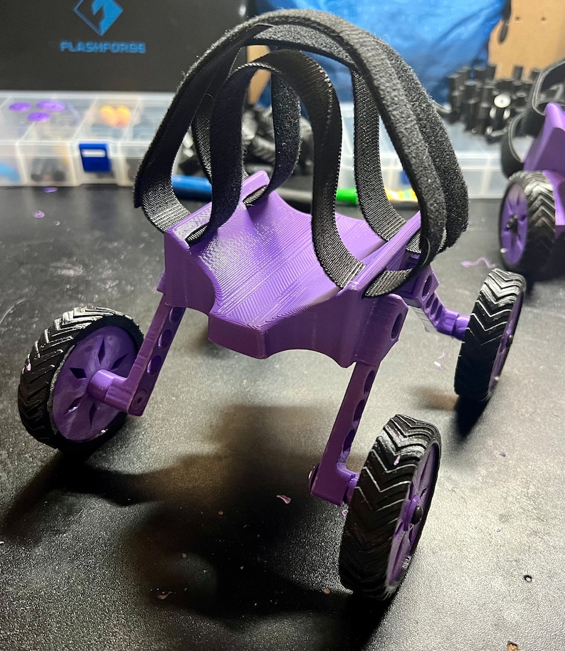 May include: A purple 3D printed, four-wheeled,  robotic walker with black straps. The walker has a unique design with a curved back and a flat base. The straps are attached to the back of the walker and are designed to help the user walk.