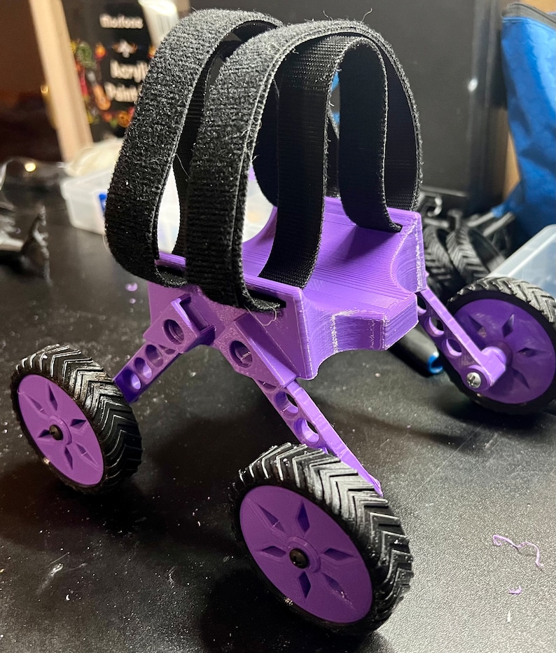 May include: A purple 3D printed toy with four wheels and black straps. The toy has a unique design and is made of plastic.