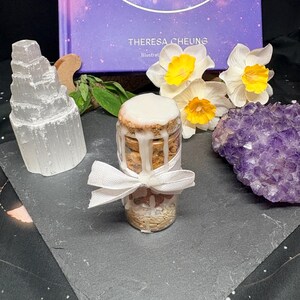 May include: A small glass jar with a white-iced top and a white ribbon tied around it. The jar is filled with small, light-colored objects. A selenite tower, a purple amethyst cluster, and a book with the word "FIX" are also in the image.