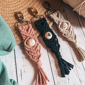 Könnte beinhalten: Drei Makramee-Schlüsselanhänger in Rosa, Schwarz und Beige. Jeder Schlüsselanhänger hat einen Holzknopf mit einem Katzen- oder Pfotenabdruck-Design und den Worten "Cat Mom". Die Schlüsselanhänger haben Metallverschlüsse und Quasten.
