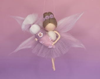May include: A handmade fairy doll with a lavender tulle skirt and delicate wings. The doll has brown hair, a white torso, and is holding a smaller doll. The fairy is adorned with a small flower and a bow. The background is a gradient of pink and purple.