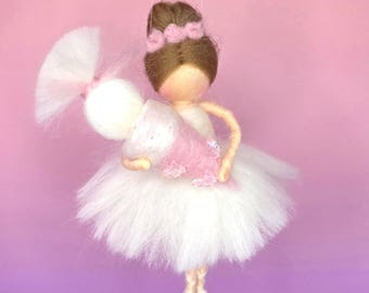 Op de afbeelding: Handgemaakte ballerina pop met een witte tutu en een roze lijfje. De pop heeft bruin haar in een knot met een roze bloemen haarband. De pop hangt aan een dun touwtje tegen een roze en paarse gradiënt achtergrond.