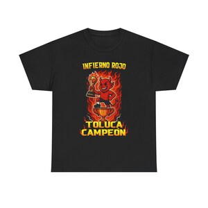 Op de afbeelding: Zwart T-shirt met een cartoonduivel die een trofee vasthoudt. De tekst "Infierno Rojo" staat erboven en "Toluca Campeon" eronder. De duivel is rood en de vlammen zijn geel en oranje.