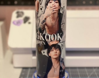 May include: A silver tumbler with a black and white image of a man with the text "GKOOK SEVEN" printed on it. The tumbler is on a silver base with a blue light.