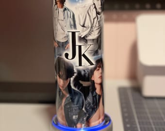 May include: A stainless steel tumbler featuring multiple images of a person in various poses, with the letters "JK" in a bold font. The design includes a light blue background with star-like accents. The tumbler is cylindrical and sits on a silver base.