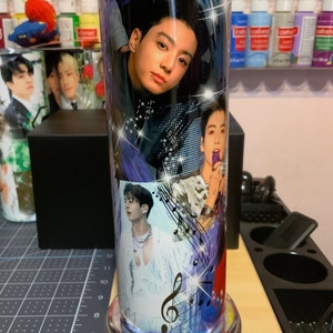 Pu&ograve; includere: Un bicchiere in acciaio inossidabile con un design colorato che presenta pi&ugrave; immagini di un membro di una band K-pop. Il design include note musicali e una chiave di violino. Il bicchiere ha una base argentata.