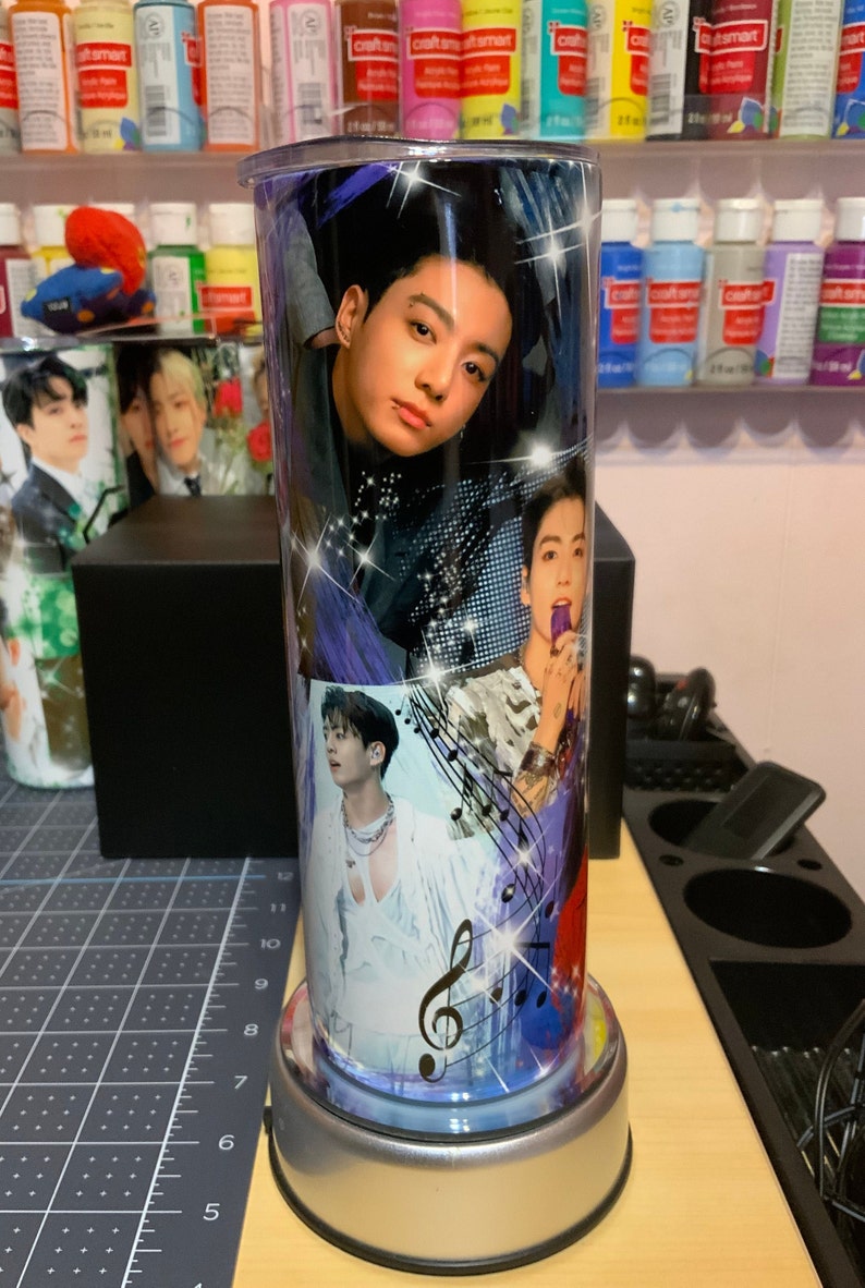 Pu&ograve; includere: Un bicchiere in acciaio inossidabile con un design colorato che presenta pi&ugrave; immagini di un membro di una band K-pop. Il design include note musicali e una chiave di violino. Il bicchiere ha una base argentata.