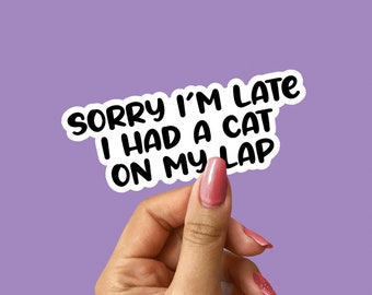 May include: A white sticker with black text that reads "Sorry I'm late I had a cat on my lap". A hand is holding the sticker against a purple background.