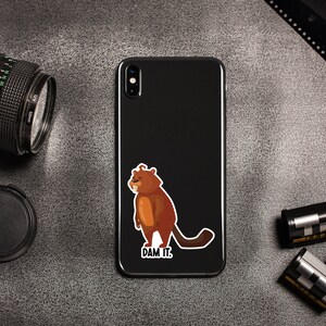 May include: A black smartphone with a brown cartoon beaver sticker on the back. The sticker says "DAM IT."