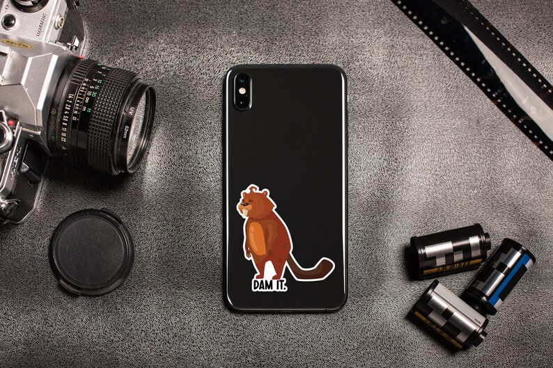 May include: A black smartphone with a brown cartoon beaver sticker on the back. The sticker says "DAM IT."