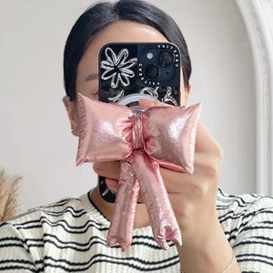 May include: A pink bow-shaped accessory is held in front of a phone with a floral design case. The person is wearing a black and white striped shirt. The bow has a shiny, metallic appearance.