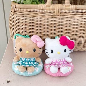 May include: Two plush Hello Kitty dolls, one tan and one white, each with a bow and dress, sit on a pink surface. A woven basket with a handle and white flowers in it is behind them. The dolls are approximately 4 inches tall.