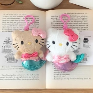 Puede incluir: Dos peluches de Hello Kitty sirena, uno beige y otro blanco, con lazos rosas y colas de sirena turquesas y moradas, están sobre un libro abierto. Un jarrón con rosas rojas, gafas y una tarjeta en blanco están en el fondo.