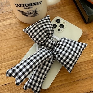 Puede incluir: Un lazo a cuadros blanco y negro con un anillo para tel&eacute;fono adjunto. El lazo est&aacute; colocado encima de un iPhone blanco con una lente de c&aacute;mara negra. El tel&eacute;fono est&aacute; sobre una superficie de madera.
