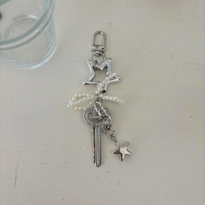 May include: Silver-toned keychain featuring a star-shaped charm, a key pendant, and a small star charm. The keychain is adorned with a string of small, white pearls. The design is elegant and suitable for keys or as a decorative accessory.