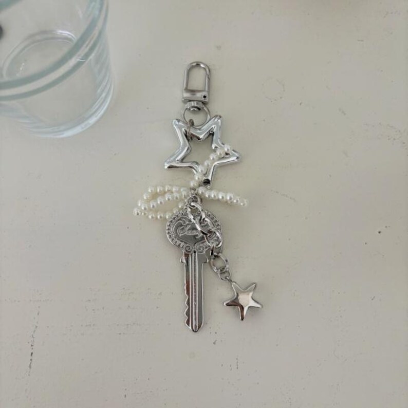May include: Silver-toned keychain featuring a star-shaped charm, a key pendant, and a small star charm. The keychain is adorned with a string of small, white pearls. The design is elegant and suitable for keys or as a decorative accessory.