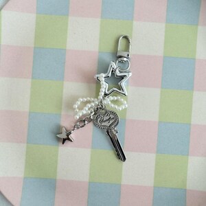 May include: Silver-toned keychain featuring a star-shaped charm, a key charm with the word "Serenity", and a small star pendant. The keychain is adorned with a string of white pearls and a silver clasp. The background is a pastel plaid pattern.