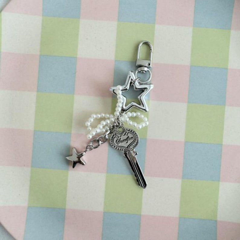 May include: Silver-toned keychain featuring a star-shaped charm, a key charm with the word "Serenity", and a small star pendant. The keychain is adorned with a string of white pearls and a silver clasp. The background is a pastel plaid pattern.