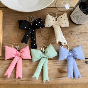 Puede incluir: Una colección de llaveros con forma de lazo en varios colores: negro, crema, rosa, verde menta y azul claro. Cada lazo presenta lunares blancos y un llavero plateado. Los llaveros están colocados sobre una superficie de madera.