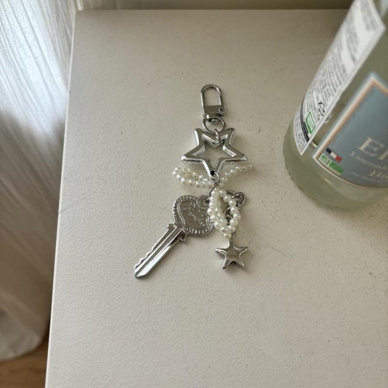May include: A silver-toned keychain featuring a key, stars, and faux pearls. The key has a decorative handle, and the stars are open-centered. A small strand of pearls hangs from the stars and key. The keychain is attached to a silver clasp.