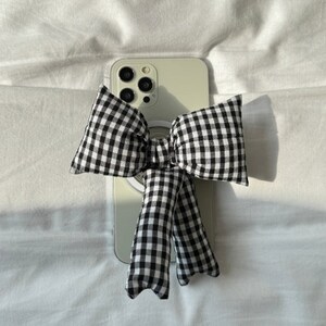 Puede incluir: Un lazo a cuadros blanco y negro con un anillo blanco unido a una funda de tel&eacute;fono blanca. La funda del tel&eacute;fono tiene una lente de c&aacute;mara negra.