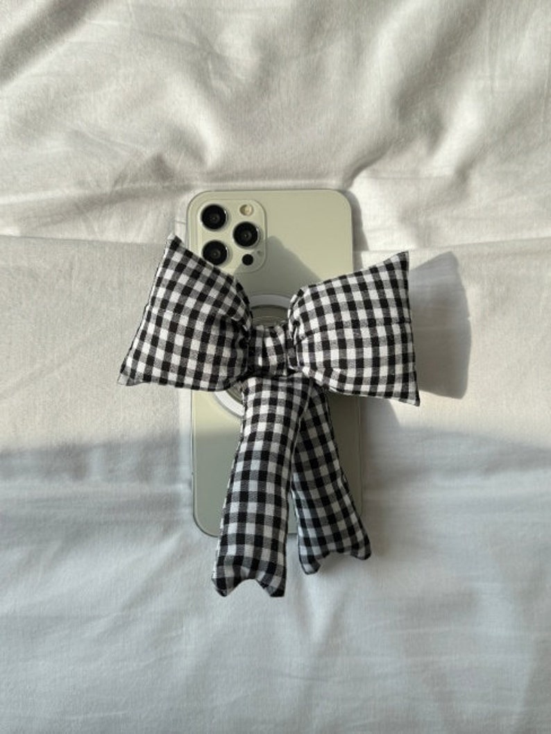 Puede incluir: Un lazo a cuadros blanco y negro con un anillo blanco unido a una funda de tel&eacute;fono blanca. La funda del tel&eacute;fono tiene una lente de c&aacute;mara negra.