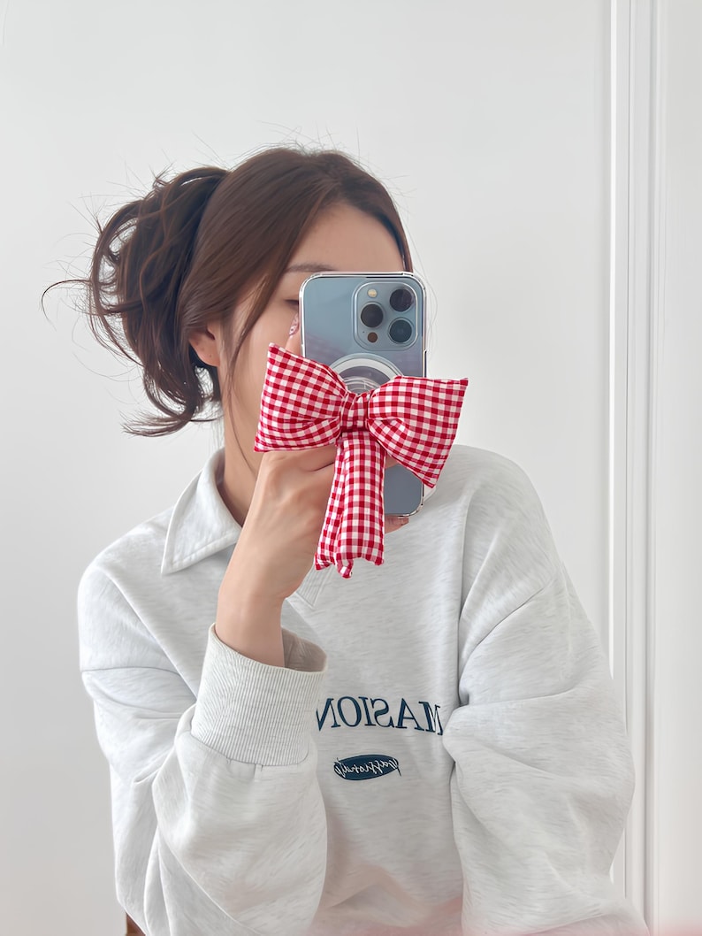 Puede incluir: Una funda de tel&eacute;fono transparente con un lazo de vichy rojo y blanco. La funda est&aacute; en un tel&eacute;fono inteligente con una lente de c&aacute;mara visible. El tel&eacute;fono est&aacute; siendo sostenido por una persona que lleva una sudadera gris claro con la palabra "MASION" impresa.