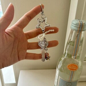 May include: Silver keychain with a star-shaped charm, a key pendant, and a small star charm. The keychain is adorned with a string of pearls. A bottle of clear liquid with a blue and orange label is in the background.