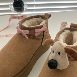 May include: Brown suede ankle boots with a plush interior and a platform sole. One boot features a pink bow, while the other has a plush dog head attached. A black cat candle sits in a glass.